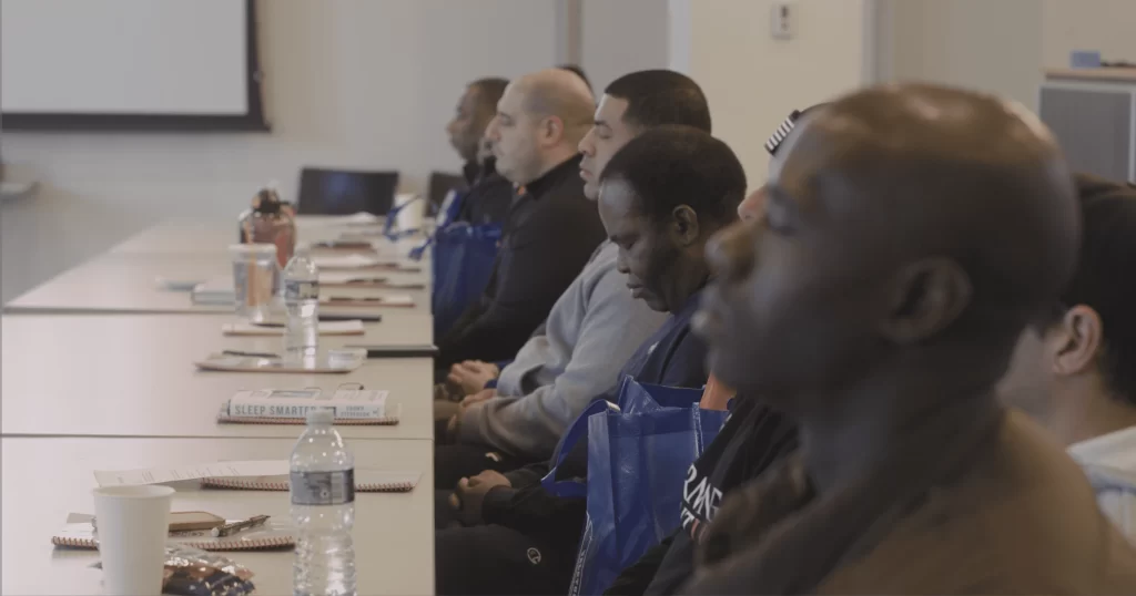 In profile, a row of adult men sit at a conference table with their eyes closed.
