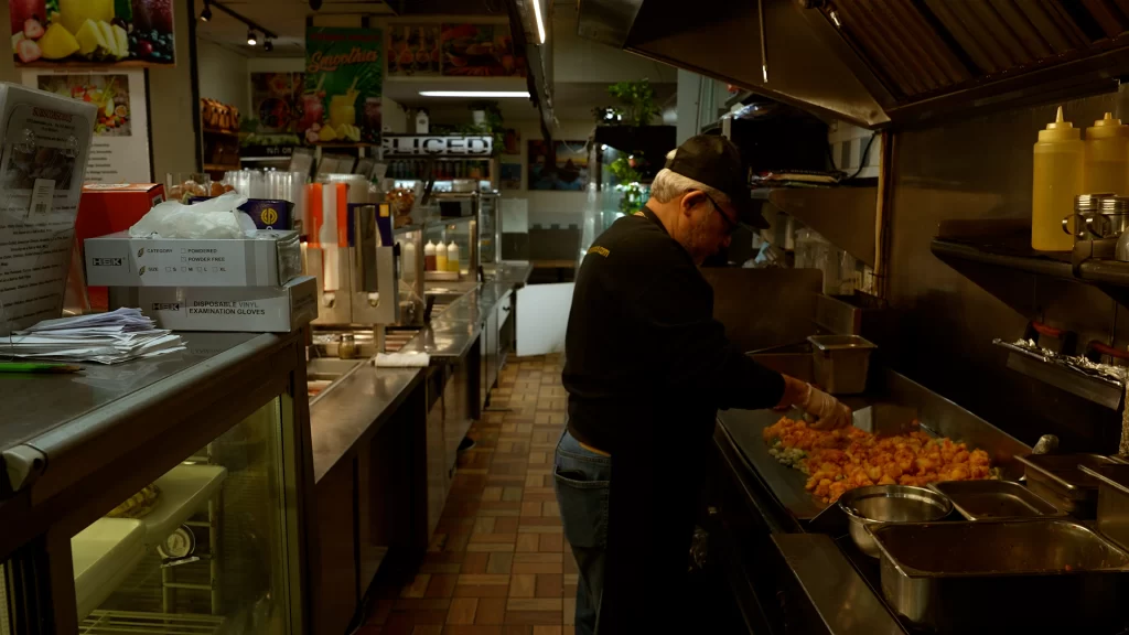 Peter Asan stands in profile in an industrial kitchen doing food prep at night. He has white hair and wears a cap and black shirt.